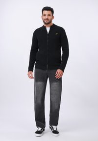 Veste zippée en tricot noir avec un tissu texturé, associée à un jean gris délavé et des baskets noir et blanc, se tenant devant un fond blanc.