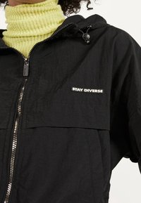 Black lightweight jacket with a textured finish, features a zip front, hood, and a patch reading "STAY DIVERSE" on the chest.