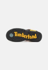Musta kumipohja, jossa on keltainen "Timberland"-logo. Pohjassa on tekstuuripatternit pidon parantamiseksi ja osia, joissa on teal-väriä.