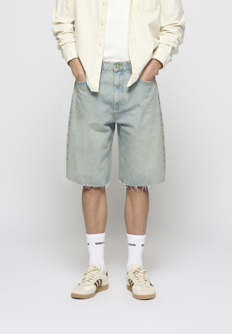 Light blue denim shorts with a loose fit, frayed hem, and visible stitching; paired with white socks and cream sneakers.