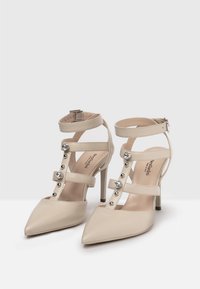 Scarpe con tacco alto beige a punta, dotate di multiple cinghie e borchie decorative in argento, realizzate in materiale sintetico liscio.