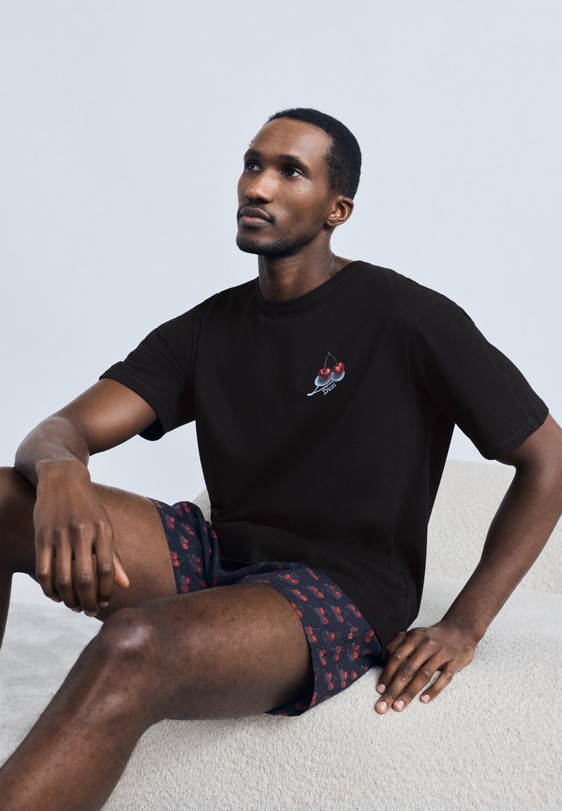 Tricou negru din bumbac cu un grafic cu cireșe; asortat cu pantaloni scurți bleumarin, cu model de cireșe. Textură fină și croială lejeră.