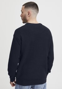 Tengerészkék bordázott kötött pulóver hosszú ujjakkal és kerek nyakkivágással, textúrázott felülettel és laza szabással. Világoskék farmernadrággal viselve.