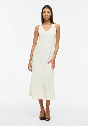 Nő ujjatlan fehér texturált midi ruhában, V-nyakkal és fekete nyitott orrú magassarkú cipőben, egyszerű fehér háttér előtt állva.