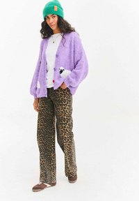 Kobieta w zielonej dzianinowej czapce, puszystym lawendowym kardiganie z naszywkami owiec, białej koszuli, spodniach w panterkę i brązowych trampkach.