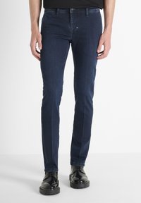 Hombre vistiendo jeans ajustados de color azul oscuro y zapatos de cordones negros pulidos, de pie contra un fondo blanco liso.