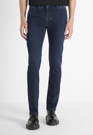 Man wearing slim-fit dark blue jeans and black polished lace-up shoes, standing against a plain white background.