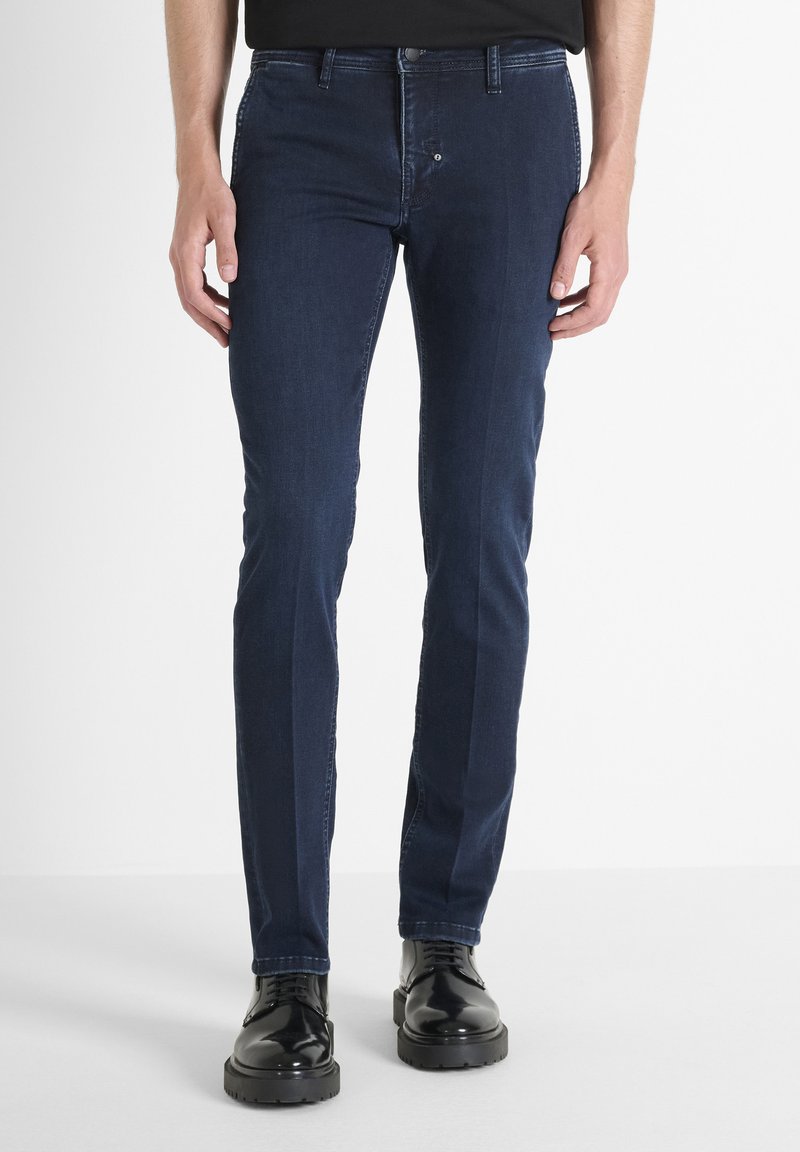 Hombre vistiendo jeans ajustados de color azul oscuro y zapatos de cordones negros pulidos, de pie contra un fondo blanco liso.