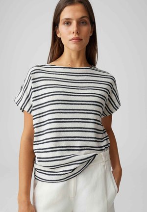 Donna con lunghi capelli castani che indossa una maglietta bianca a maniche corte con strisce orizzontali nere e pantaloni bianchi, in piedi davanti a uno sfondo grigio.