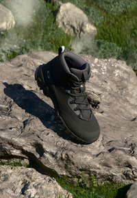 Crne planinarske čizme s robusnim Vibram potplatom odmaraju se na velikom kamenu na otvorenom uz travu i kamenčiće u pozadini.