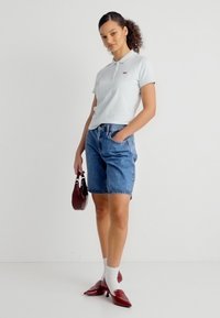 Γυναίκα που φοράει λευκό πόλο πουκάμισο, τζιν σορτς, λευκές κάλτσες, κόκκινες γόβες και κρατάει μια μικρή κόκκινη τσάντα χειρός.