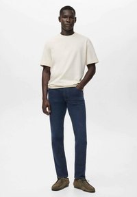Hombre de pie con la mano izquierda en el bolsillo, vistiendo una camisa corta de manga crema, jeans azul oscuro y zapatos marrones con cordones sobre un fondo blanco.