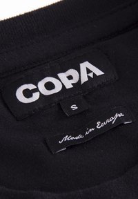 Zwarte stoffen label met witte "COPA" logo en maat "S." Hieronder staat in kleinere letters "Made in Europe" in cursief. Zachte textuur, reguliere vorm.