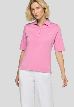 Frau mit kurzen blonden Haaren, die ein rosa kurzärmeliges Poloshirt und weiße Hose trägt, steht vor einem hellgrauen Hintergrund.