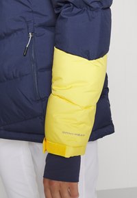 Giacca a vento con parte superiore blu navy e manica gialla brillante. Tessuto testurizzato con marchio "OMNI-HEAT" sul polsino. Dettaglio con tasca con zip.