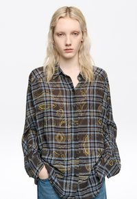 Camisa de manga larga con cuello en marrón oscuro y cuadro azul con acentos amarillos, que presenta un cierre de botones en la parte delantera y un corte holgado.