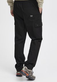 Sorte cargo bukser med elastisk talje, to sidelommer og et synligt logo-patch på højre baglomme. Holdbar stoftekstur.