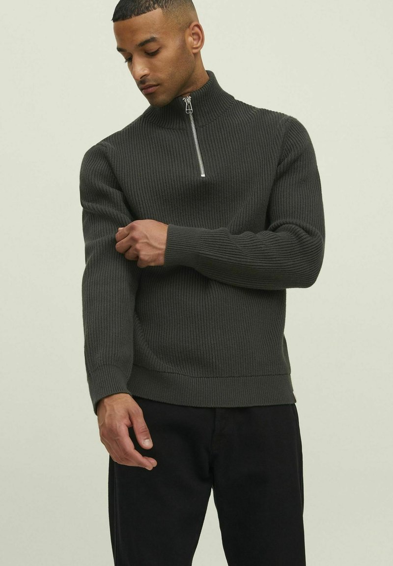 Dunkelgrüner gerippter Pullover mit hohem Kragen, silbernem Reißverschluss und langen Ärmeln. Das Material scheint strukturiert und hat ein tailliertes Design.