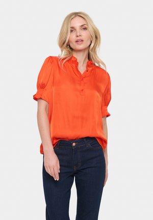Blusa arancione brillante a maniche corte con scollo arricciato e chiusura a bottoni; abbinata a pantaloni in denim blu scuro. Tessuto liscio e lucido.