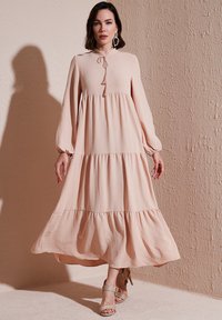 Lang, lagdelt kjole i lys ferskenfarvet stof med høj hals, lange ærmer og snøredetalje. Kombineret med beige strop hæle.