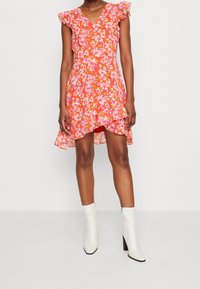Blumenkleid mit einem lebhaften orangefarbenen Grund, Rüschensäumen, V-Ausschnitt, taillierter Taille und ausgestelltem Saum; kombiniert mit weißen Absatzstiefeln.