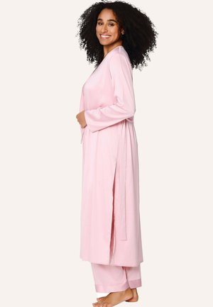 Usmievavá žena s kučeravými vlasmi v svetloružovom dlhom župane a zodpovedajúcich nohaviciach, stojaca bosá pred jednoduchým pozadím.