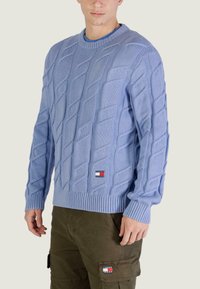 Pull en tricot torsadé bleu clair avec poignets et ourlet côtelés, orné d'un petit patch logo rouge, blanc et bleu sur le bas devant.