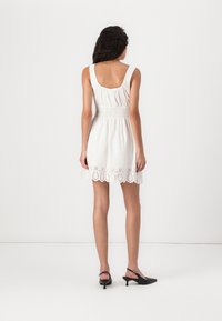 White sleeveless dress with a smocked bodice and scalloped lace hem; paired with black slingback heeled shoes. Smooth texture, casual design.