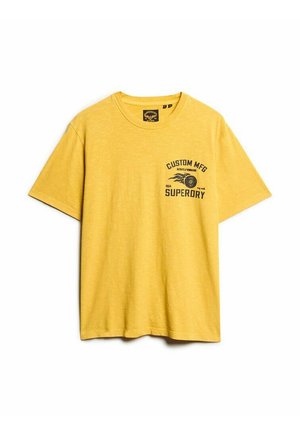 Żółta bawełniana koszulka z okrągłym dekoltem i krótkimi rękawami. Na przodzie znajduje się grafika z tekstem "CUSTOM MFG" oraz "SUPERDRY" w kolorze czarnym.