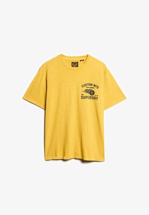 Żółta bawełniana koszulka z okrągłym dekoltem i krótkimi rękawami. Na przodzie znajduje się grafika z tekstem "CUSTOM MFG" oraz "SUPERDRY" w kolorze czarnym.