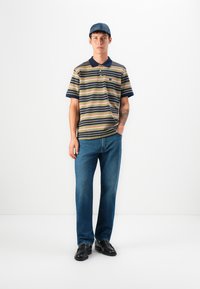 Polo rayé beige, bleu et rouge à manches courtes ; associé à un jean bleu et des chaussures noires ; design décontracté avec un col.
