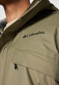 Homme portant une veste Columbia vert olive avec logo visible sur la poitrine et rabat de poche incliné avec tirette de fermeture éclair noire.