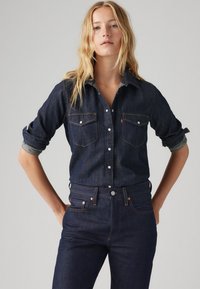 Camisa de ganga escura com frente abotoada, dois bolsos no peito e mangas dobradas. Apresenta costura contrastante e um design de colarinho clássico.