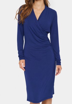 Vrouw draagt een marineblauwe wikkeljurk met lange mouwen en knie lengte, met geplooide details aan de zijkant, poserend tegen een effen achtergrond.