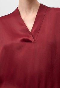 Blusa in seta bordeaux con scollatura a V profonda e texture morbida, caratterizzata da linee pulite e design minimalista. Nessun motivo visibile o decorazioni.
