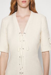 Robe couleur crème en tissu texturé avec un décolleté en V profond, manches courtes, et un devant lacé avec des œillets métalliques et des liens en ruban.