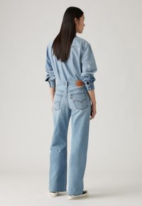 Light blue denim outfit showcasing a relaxed fit with long sleeves, rolled cuffs, and a back patch label. Flat, wide legs tapering at the hem.