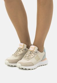 Zapatillas deportivas beige con parte superior sintética texturizada, acentos en azul y durazno, diseño con cordones y suela blanca gruesa con tracción.