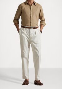 Camicia button-up marrone chiaro, pantaloni bianchi plissettati, cintura in pelle marrone, e scarpe marrone scuro, caratterizzate da un design elegante e una vestibilità su misura.