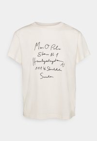 Camiseta de algodón color crema con un corte relajado. Presenta texto manuscrito en negro que incluye el nombre de la marca y la dirección. Mangas cortas y escote redondo.