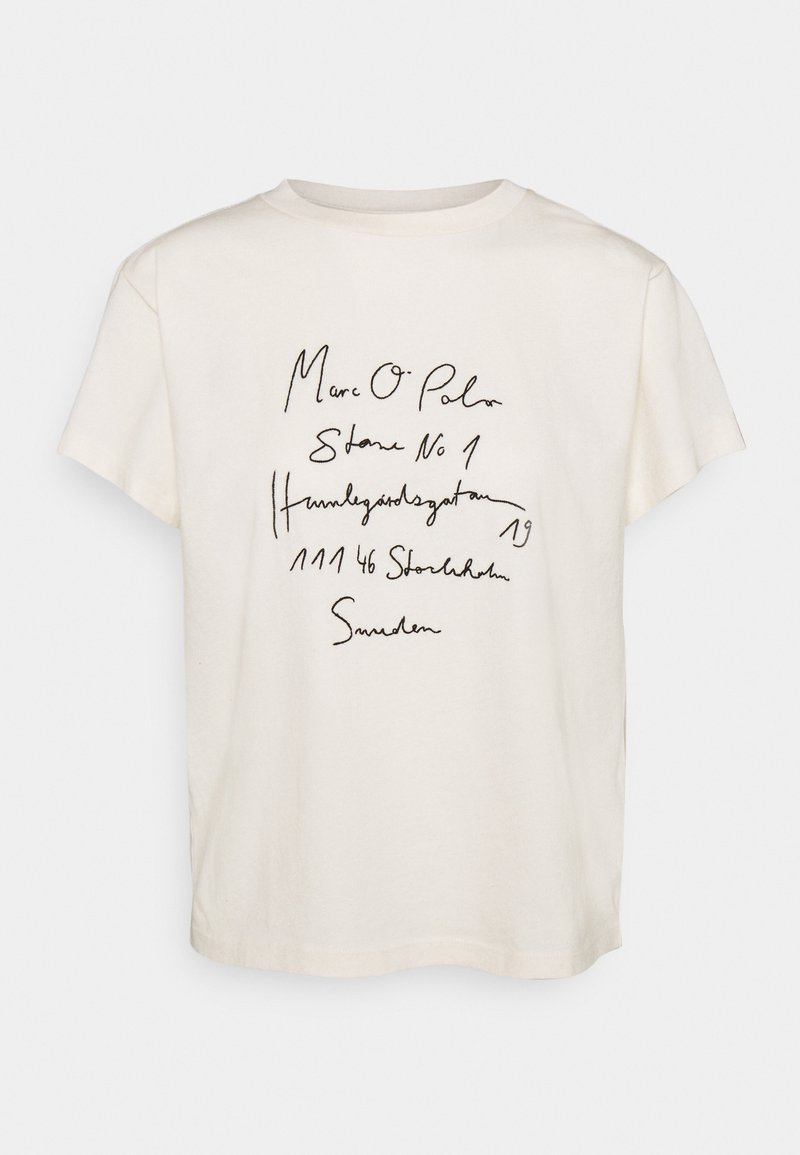 Camiseta de algodón color crema con un corte relajado. Presenta texto manuscrito en negro que incluye el nombre de la marca y la dirección. Mangas cortas y escote redondo.