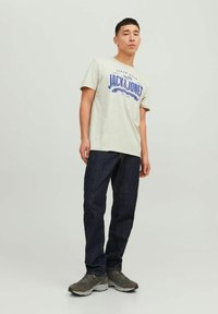 Grå bomulds t-shirt med blåt grafisk print, parret med mørkeblå denim jeans. Modellen har grå træningssko på. Enkel, afslappet design.