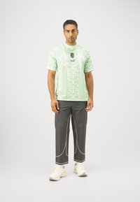 Camiseta verde con estampado y mangas cortas, combinada con pantalones grises con acentos de costura blanca. El calzado son zapatillas cremosas.