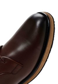 Scarpa in pelle marrone con punta arrotondata, texture liscia e suola di un marrone chiaro a contrasto. Presenta dettagli cuciti e design con lacci.