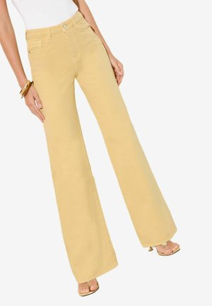 Kvinde iført beige vidde bukser, hvid top, gyldne armbånd og gyldne åbne hæle, stående mod en ensfarvet baggrund.