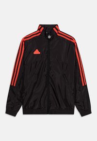 Musta urheilujakku, jossa on punainen kolmiraidallinen muotoilu, korkea kaulus, vetoketjukiinnitys, joustavat hihansuut ja taskut. Valmistettu kiiltävästä, sileästä kankaasta.