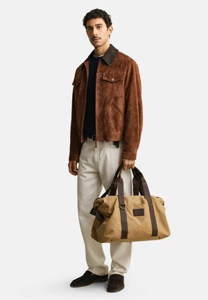 Homme portant une veste en daim marron et un pantalon blanc, tenant un sac de voyage en toile beige avec des sangles marron foncé, debout devant un fond uni.