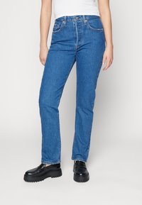 Personne portant un jean bleu taille haute et des mocassins à semelle épaisse noirs, se tenant debout avec les mains détendues le long du corps.