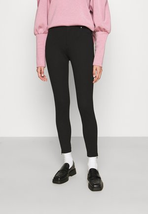 Person, die schwarze Skinny-Hosen, weiße Socken, schwarze Loafers und ein rosa langärmeliges Oberteil mit Puffärmeln trägt, steht auf einem grauen Boden.