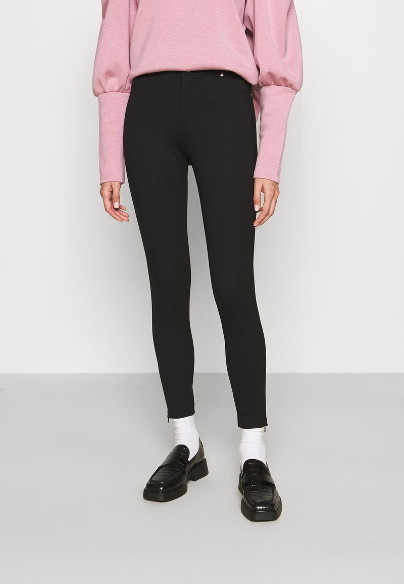 Personne portant un pantalon skinny noir, des chaussettes blanches, des mocassins noirs et un haut rose à manches longues bouffantes, debout sur un sol gris.